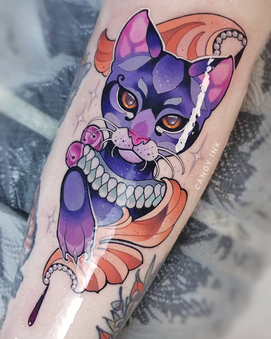 a freshly done neo traditional tattoo of a purple cat wearing a pearl necklace with a red jewel on its arm.  it is on someone's inner forearm.  the tattoo has highlights in a lighter purple.