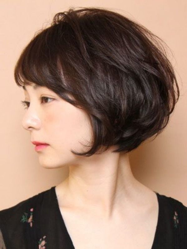 a young asian woman with short, dark brown hair styled in a choppy bob with bangs, is wearing a black dress with a floral print. she is looking down and has a gentle, delicate expression. the image is closeup, focusing on her face and hair. the background is a soft, light color.  she is wearing a black dress with a floral print that covers her shoulders. the image is a closeup portrait, focusing on her face and hair. the background is a soft, light color. the photo gives off an elegant, sophisticated vibe.