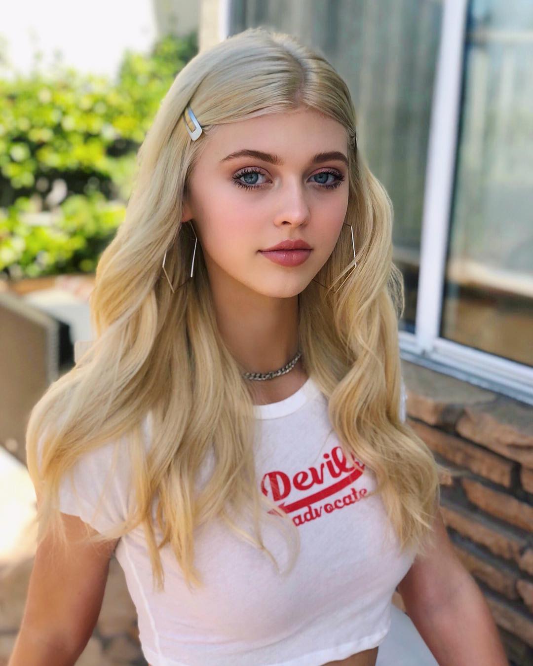 a portrait shot of a young woman with blonde hair, blue eyes and fair skin wearing a white tshirt with the words "devil advocate" written in red on it. she has a silver hair clip in her hair and is wearing square earrings and a silver chain necklace.