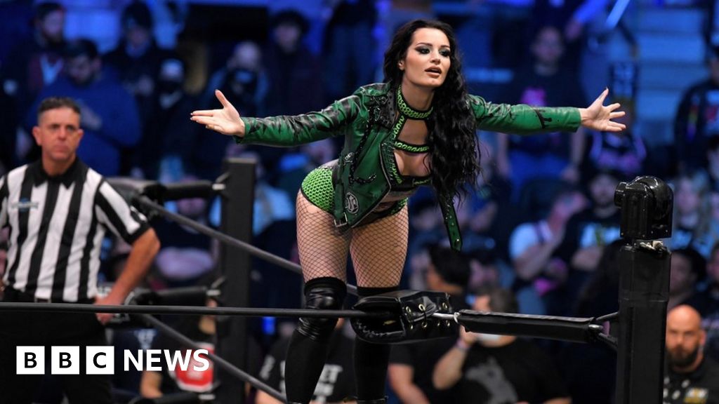the image shows a female professional wrestler, likely from aew judging by the style, standing in a wrestling ring.  she's wearing a green leather jacket, a matching green and black top, black fishnet stockings, and black kneehigh boots.  her arms are outstretched, possibly in a gesture of defiance or victory. a referee in a black and white striped shirt stands in the background. the background is blurry but shows a large crowd in an arena setting. the bbc news logo is superimposed on the bottom left corner of the image. the overall impression is one of action and excitement within the context of a professional wrestling match.