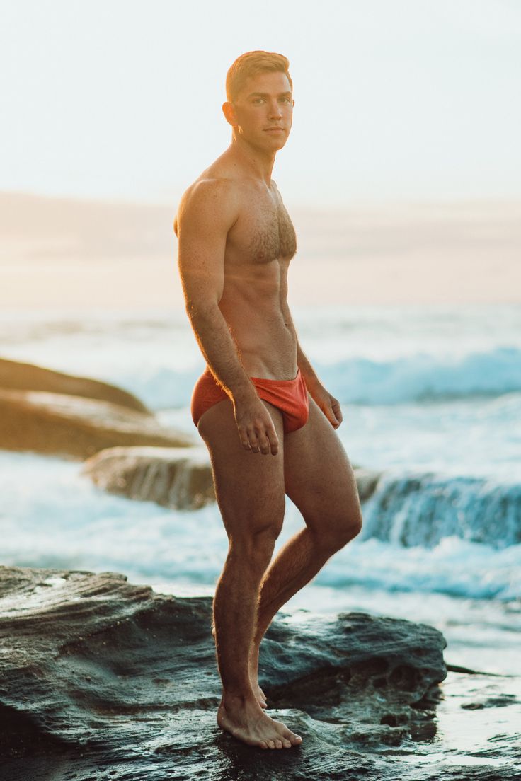 a young, caucasian male model is standing on a rocky beach, wearing red swim trunks and looking at the camera. the ocean waves lap at the rocks behind him. the sun is setting or rising, casting a warm glow over the scene. the model is fit and toned, showing off his physique. he is wearing a small amount of chest hair and appears to have a six pack. his hair is a light brown, and he has a determined look on his face. this image captures the beauty of the beach and the attractiveness of the model.