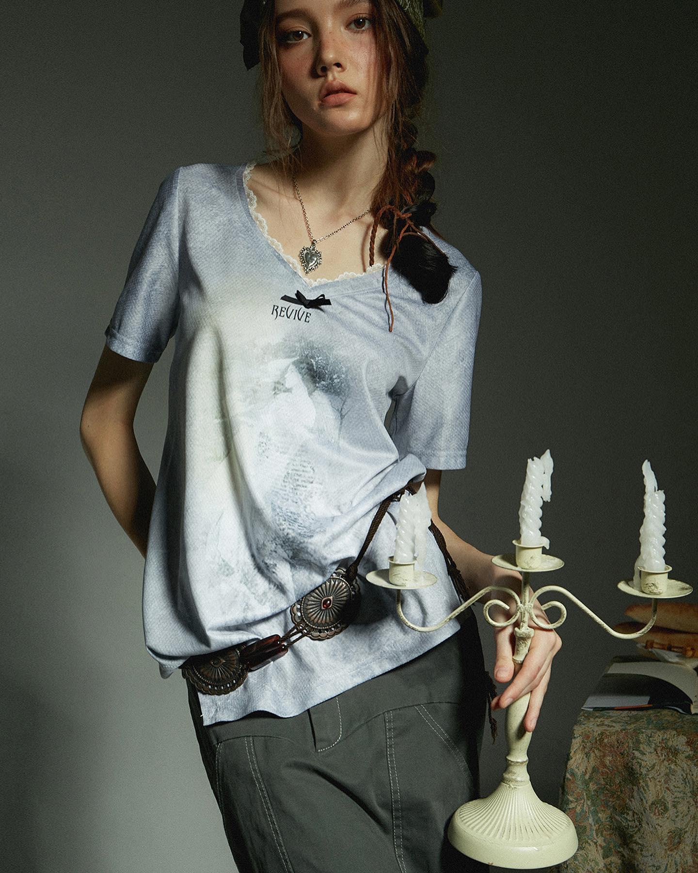 a young woman with long braided hair, wearing a grey and light blue oversized vneck tshirt featuring an abstract print and the word "revive," is shown in a studio shot. she is also wearing a vintagestyle belt with a large circular buckle and a heartshaped necklace. she holds a pale green, ornate candlestick holder with three unlit candles. the background is dark and moody, enhancing the overall style which is bohemian, grunge, and slightly rebellious. the image focuses on fashion and the model's styling.