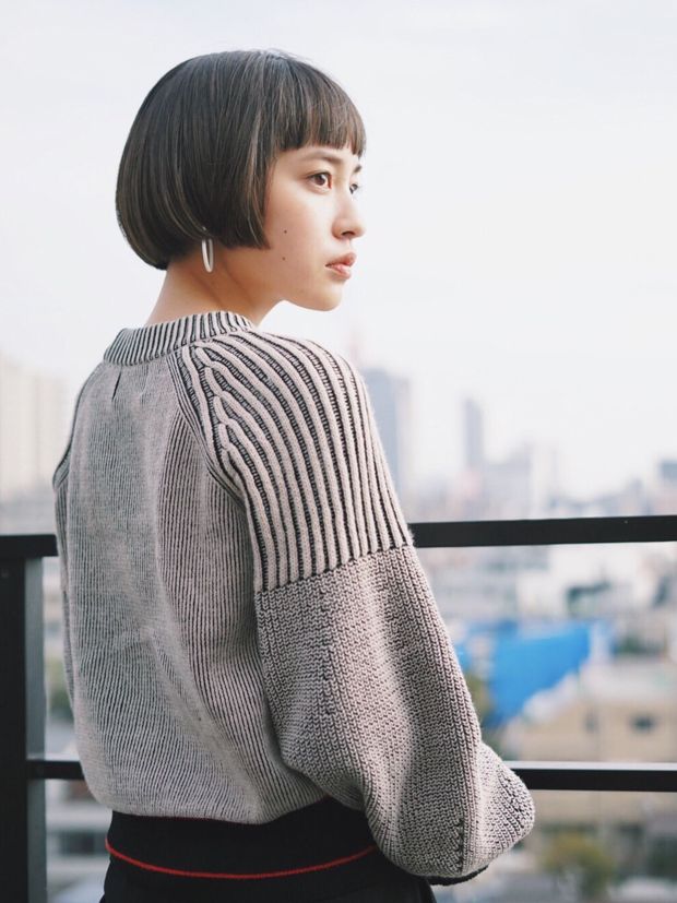 a young woman in a grey sweater stands by a balcony railing, looking out over a cityscape. she has short, black hair with bangs and wears a grey sweater that appears to be knitted. she is standing with her back to the camera, gazing towards the distance.