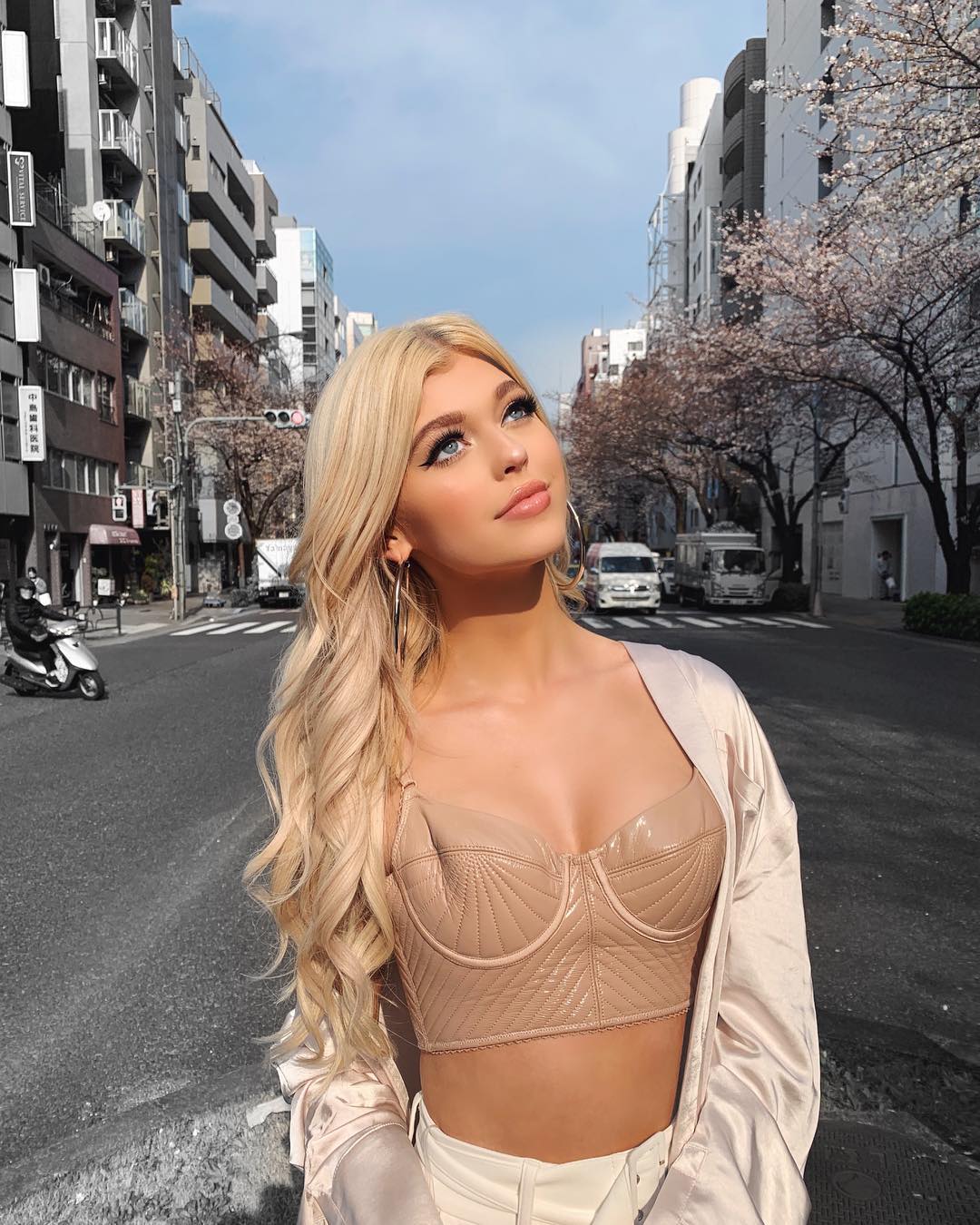 a young blonde woman, loren gray, stands on a city street, looking up towards the sky. she is wearing a nudecolored crop top, white pants, and a lightcolored jacket. her blonde hair is styled in loose waves, and she is wearing hoop earrings. the background features buildings, cherry blossoms, and a street scene with vehicles and pedestrians. the overall impression is of a fashionable woman enjoying a sunny day in an urban setting.