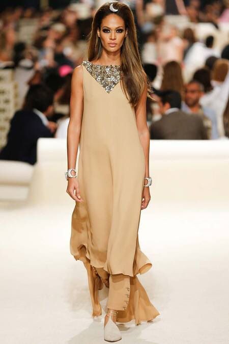 a beautiful female model walks down a runway in a long, beige, flowy dress, accessorized with a sparkly neckline, bracelets, and heels. the model is posing confidently for the camera, showcasing the high fashion of the design.