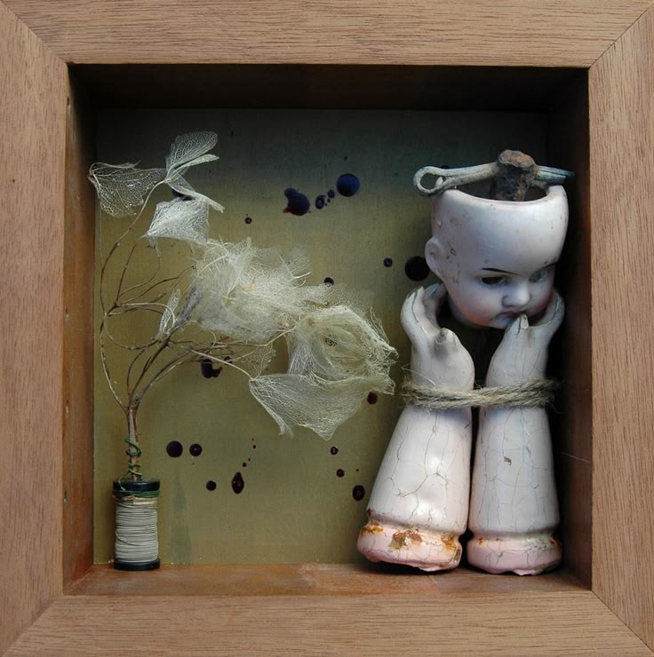 a shadow box containing an assemblage of found objects. the primary focus is a broken antique doll head positioned atop the doll's arms, which are bound together with rope. inside the doll's head is a metal object. to the left, a dried flower arrangement extends from a thread spool. dark splatters appear on the background. the box is framed with light wood.