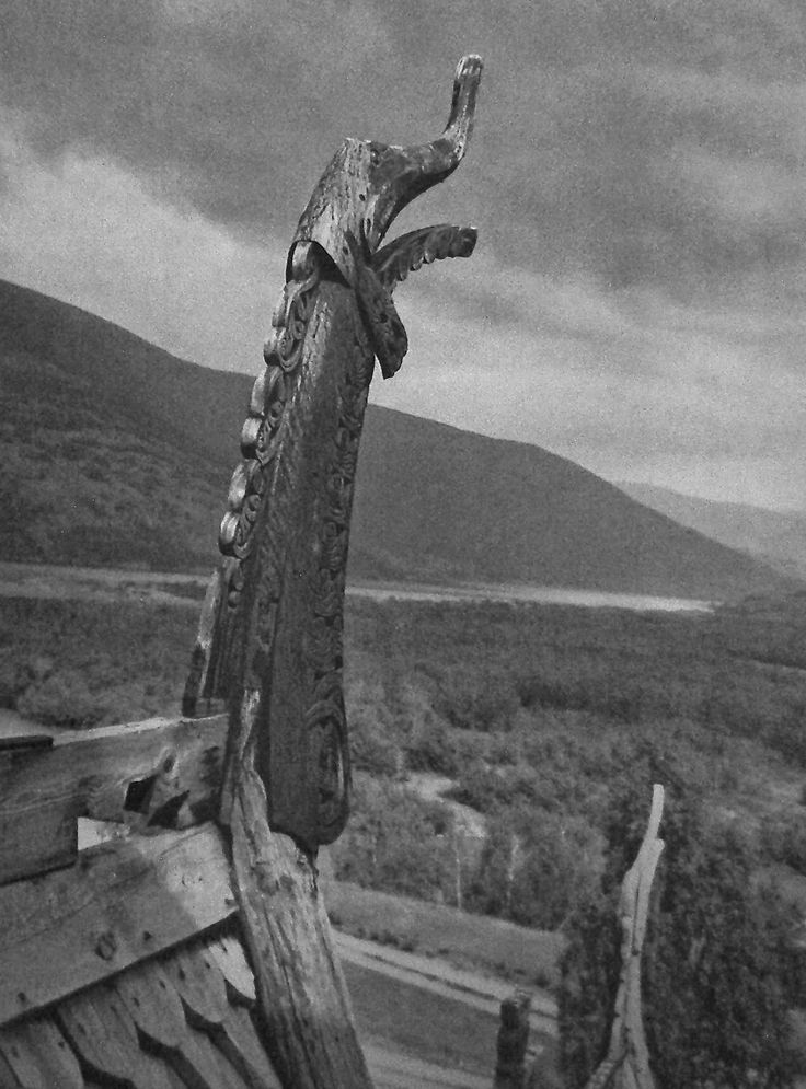 this black and white photograph shows the intricately carved dragon head of a viking ship's stern, set against a backdrop of mountains, a valley, and cloudy skies. the detailed wood carving stands out prominently, with its intricate design and craftsmanship. the landscape behind the ship adds depth and context to the historical artifact.
