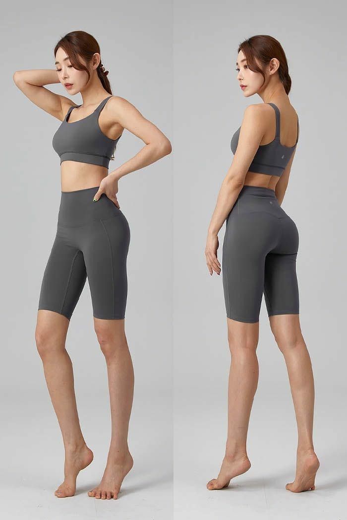 a young woman is posing in a grey sports bra and shorts. she is standing with her feet together and her arms at her sides. her hair is down and she is looking at the camera. she looks athletic and healthy.  the image is shot from a side angle. the background is a plain white wall.