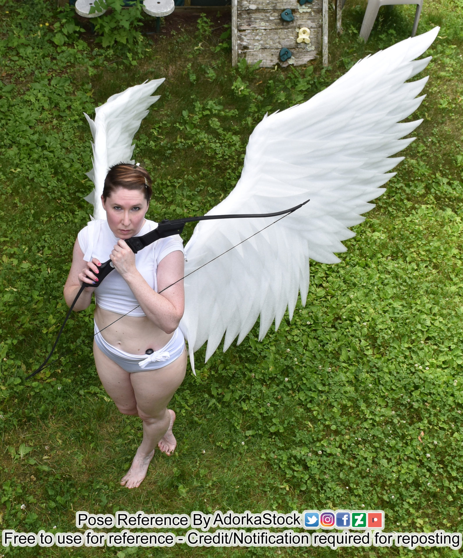 a woman wearing white angel wings and a white crop top and bottoms holds a bow and arrow. she is standing in green grass with her wings spread out behind her.