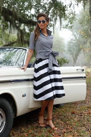 a woman in a black and white striped skirt and a checkered top, standing next to a classic car and posing for the camera. she is wearing sunglasses and smiling.