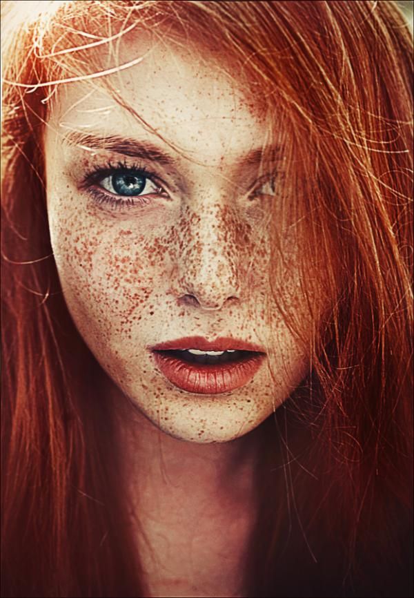 the image shows a closeup portrait of a young woman with striking features. she has vibrant red hair, fair skin covered in freckles, and piercing blue eyes. the focus is on her face, capturing her natural beauty.
