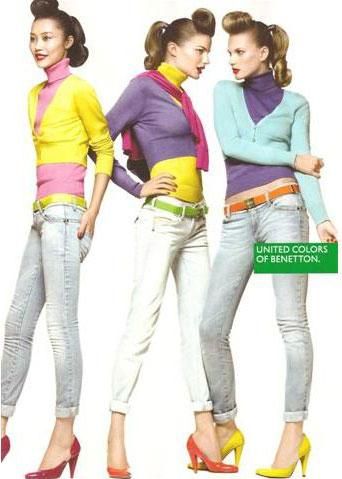 three women are posing in an advertisement for united colors of benetton. they are wearing brightly colored sweaters and turtlenecks, jeans, belts, and high heels. the woman on the left is wearing a yellow sweater with a pink turtleneck and red high heels. the woman in the middle is wearing a purple sweater with a yellow stripe and orange high heels. the woman on the right is wearing a blue sweater with a purple turtleneck and yellow high heels. they all have their hair styled in a similar way, with a small bun on top of their heads. a green rectangle with the words "united colors of benetton" is on the right side of the image.