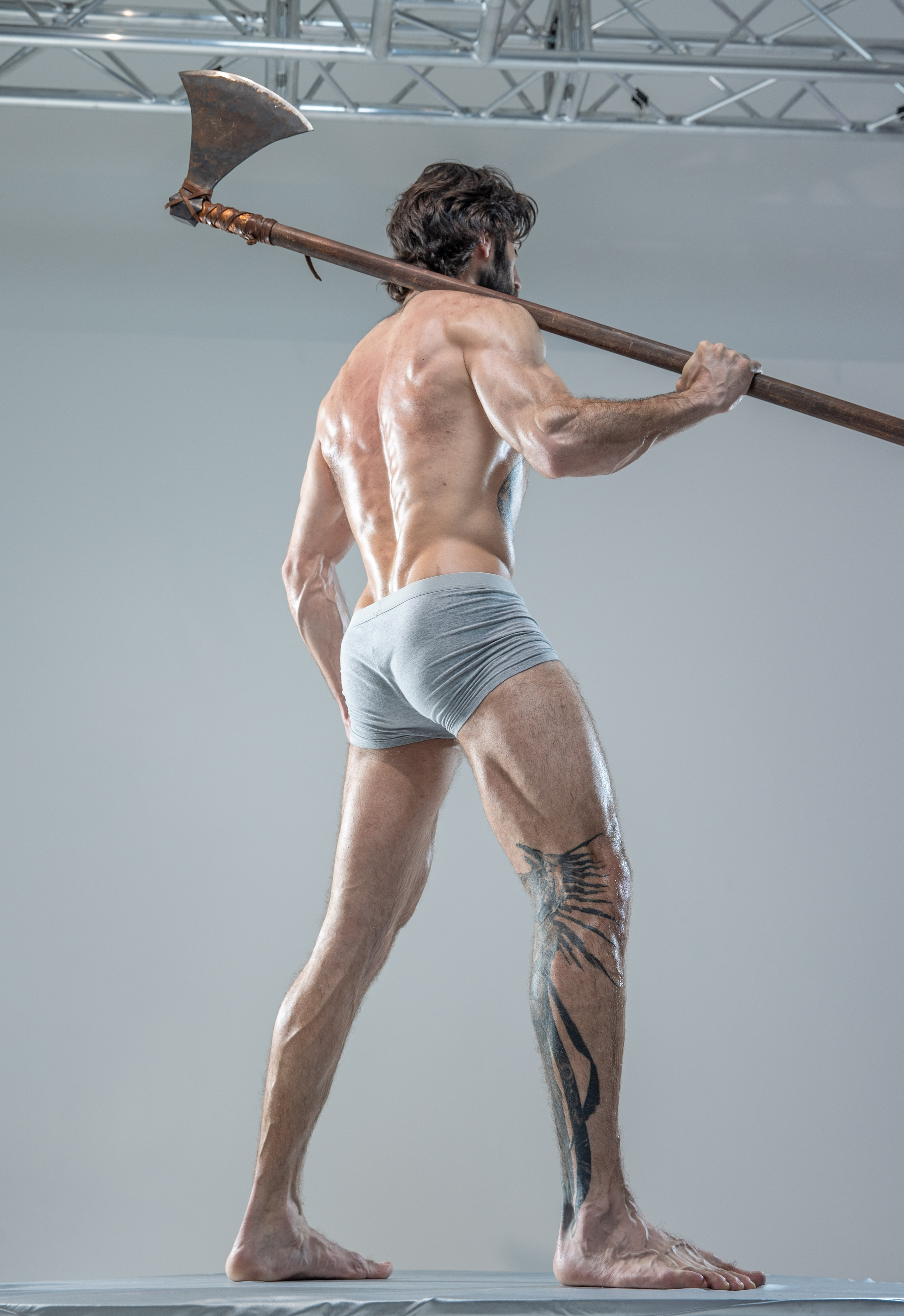 a muscular man with sweat glistening on his skin stands with his back to the camera. he holds a large, worn axe across his shoulders. his pose emphasizes the definition of his back muscles. he is wearing only gray underwear and stands in a studio setting with artificial lighting. the perspective of the image is from a low angle, highlighting the power and strength of his stance.