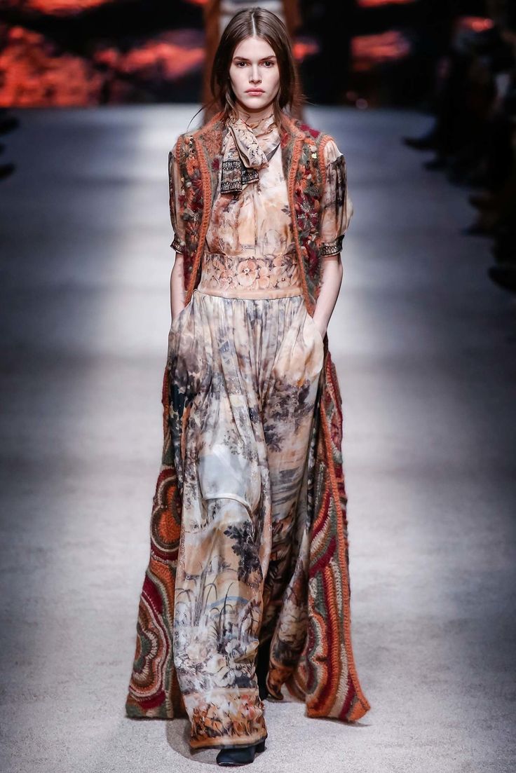 a model walks the runway in a long, flowing outfit. the ensemble features a printed, loosefitting pantsuit with a landscapelike pattern in muted earth tones.  over this, she wears a cropped, intricately textured vest in a mix of browns and reds, seemingly crocheted or knitted.  a silk scarf is casually tied around her neck. her hair is styled in a sleek, centreparted look. the background is blurred, focusing attention on the model and her attire. the overall style is bohemian or ethnicinspired, suggesting autumn or winter fashion.