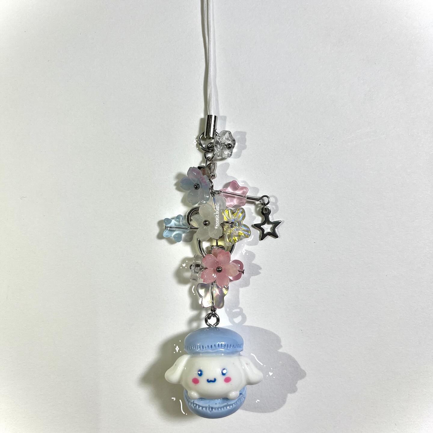 a closeup shot of a keychain charm. the charm consists of a cinnamoroll figure encased in a blue macaron shape, connected to a string of pastelcolored flowershaped beads, and a silver star charm. the keychain has a white string for attachment. the background is a plain white surface.