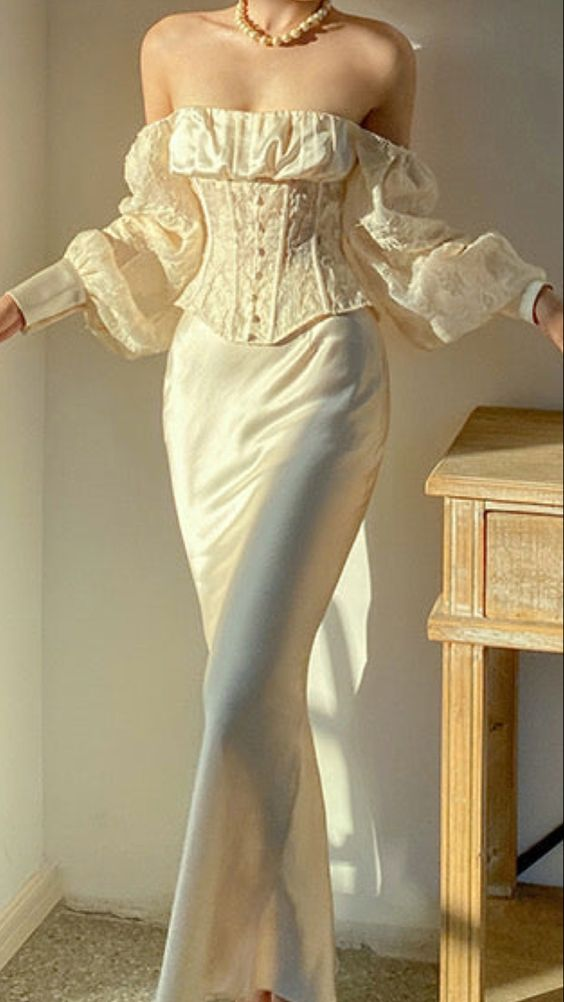 a woman wearing a champagnecolored offtheshoulder corset top with long sleeves and a matching silk skirt. the top features intricate lace detailing and a boned bodice. the skirt is long and flowing, with a slight sheen. the woman is standing in a room with sunlight streaming in. the image evokes a sense of elegance, femininity, and romance.