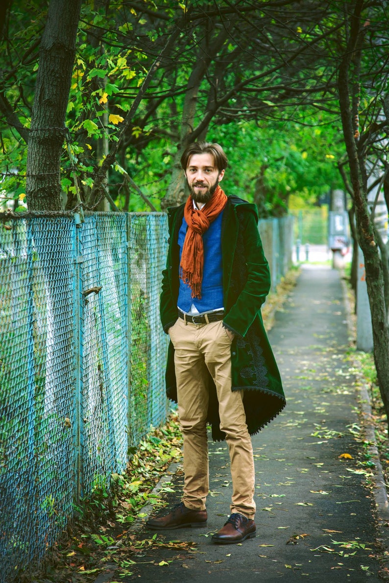 a full shot of a man standing on a walkway between trees. he is wearing a dark green long coat, a blue vest, khaki pants, brown shoes, and an orange scarf. the walkway is lined with trees and a chainlink fence is seen in the background. the leaves on the trees suggest it is autumn.