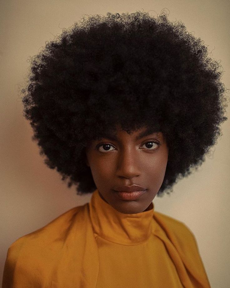 a portrait of a black woman with a large afro, wearing a yellow highcollared shirt. she is looking directly at the camera. the background is a neutral color.