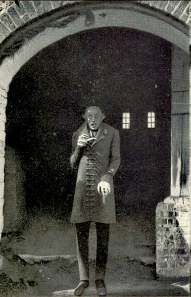 a grainy black and white photo of a man in a long coat and hat standing in a brick archway. he has a gaunt face and holds what appears to be a small object in his hand. the lighting is dim, creating a shadowy and slightly sinister atmosphere. the background features an open doorway with two small windows, suggesting an urban or possibly institutional setting.