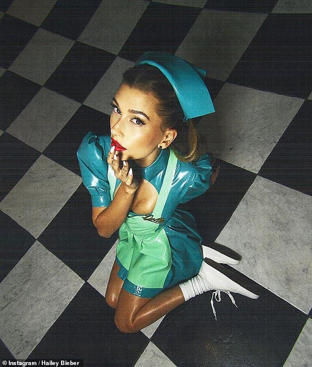 a highangle, full shot of hailey bieber kneeling on a black and white checkered floor. she is dressed in a teal and light green latex nurse costume, complete with a matching cap and white socks and shoes. she is blowing a kiss at the camera, and her makeup is bold with red lipstick. the overall aesthetic is retro and glamorous. the image is likely from a photoshoot.