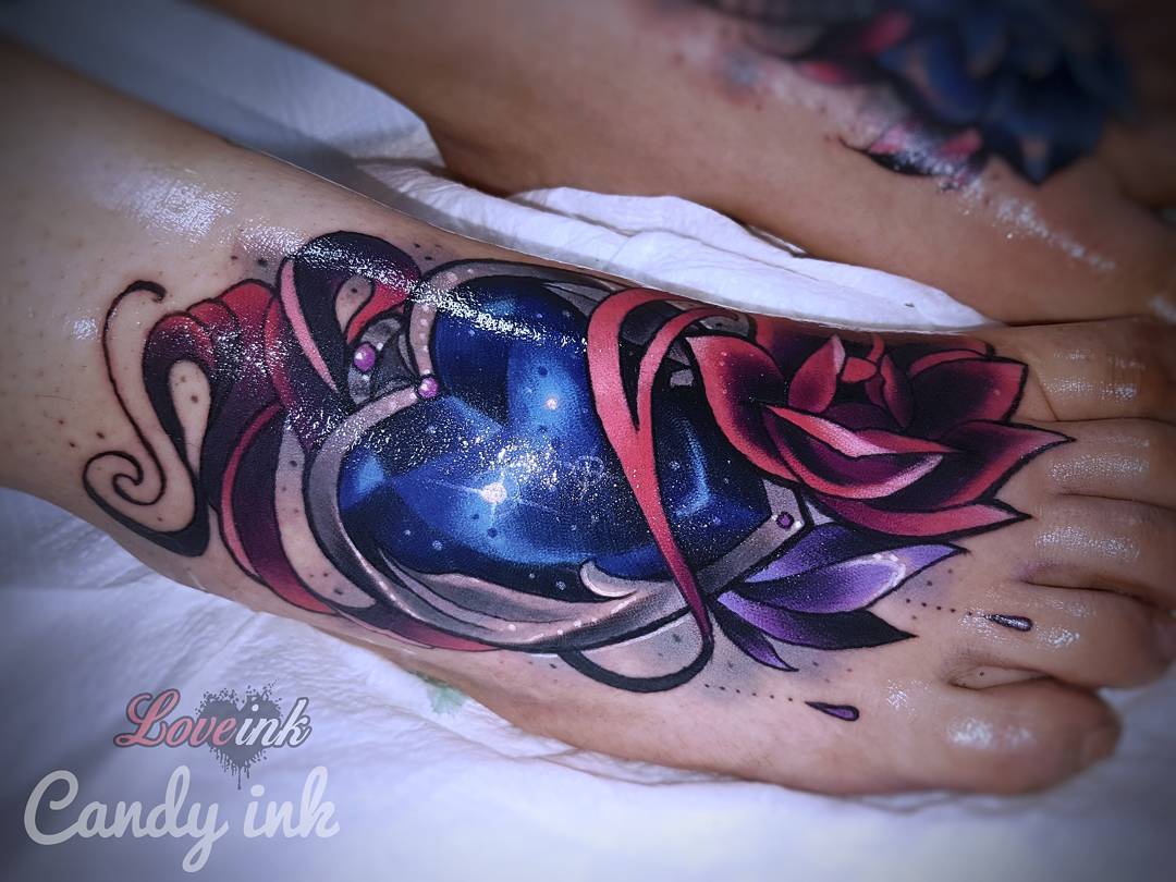 a colorful tattoo of a galaxy heart with a jewel accent on top is inked on the top of a person's foot, wrapping around the ankle. the heart is framed with a purple rose on the right and a maroon ribbon on the left.  the tattoo is clean and the colors are vibrant.