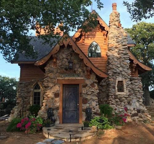 the image shows a charming fairytalelike cottage with a stone facade, wooden accents, and an ornamental roof. a tall stone chimney stands on one side, while flowers and plants surround the entrance, guarded by small lion statues. a stone walkway leads to the dark blue, gothicstyle door. the house is nestled among trees, adding to its whimsical appeal.