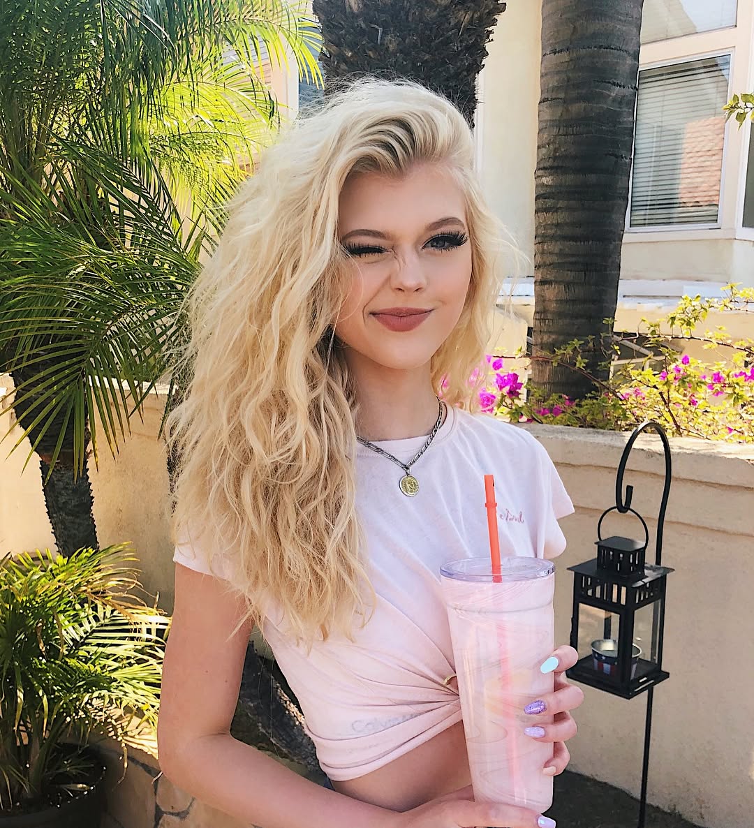 a young woman with blonde wavy hair is winking and holding a pink drink with a straw. she's wearing a pink tshirt and a necklace. the background features a palm tree, bougainvillea, and a lantern. the setting appears to be outdoors.