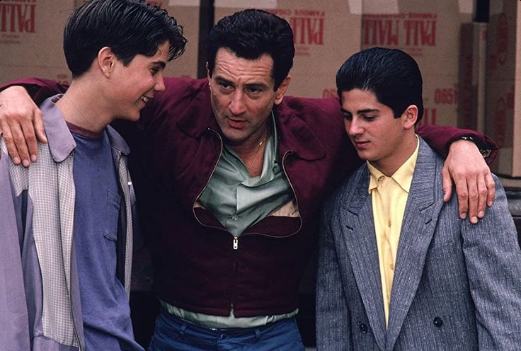 a scene from the movie a bronx tale featuring robert de niro flanked by actors lillo brancato and francis capra. in the background are boxes of palla mall cigarettes. de niro has his arms around the two younger actors. all three are wearing jackets.