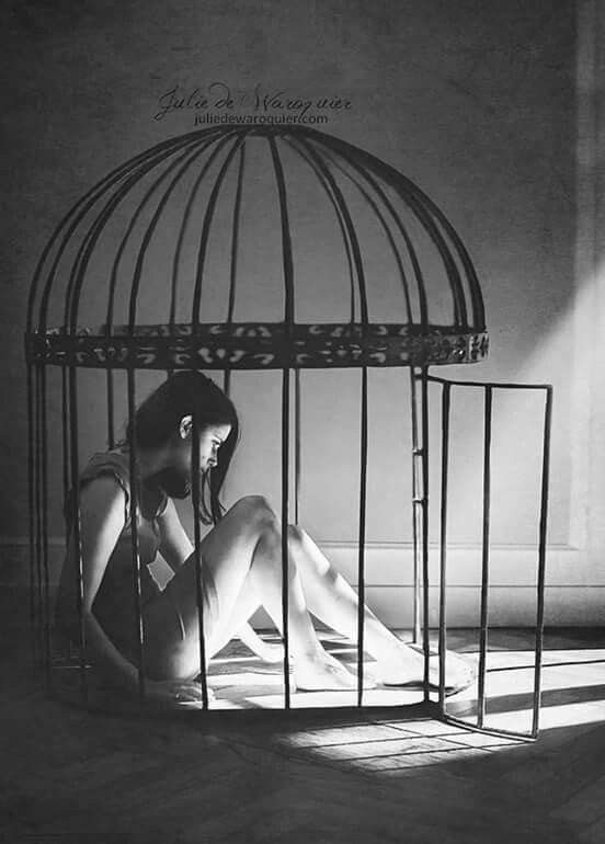 a black and white photo of a woman trapped inside a cage. the woman is sitting on the floor with her back to the wall and her legs stretched out in front of her. the cage is made of metal bars and is large enough to fit the woman inside. the image is dark and shadowy, with light coming in from the right side of the frame. the woman's facial expression is one of sadness and despair. the image suggests a feeling of captivity and restriction.