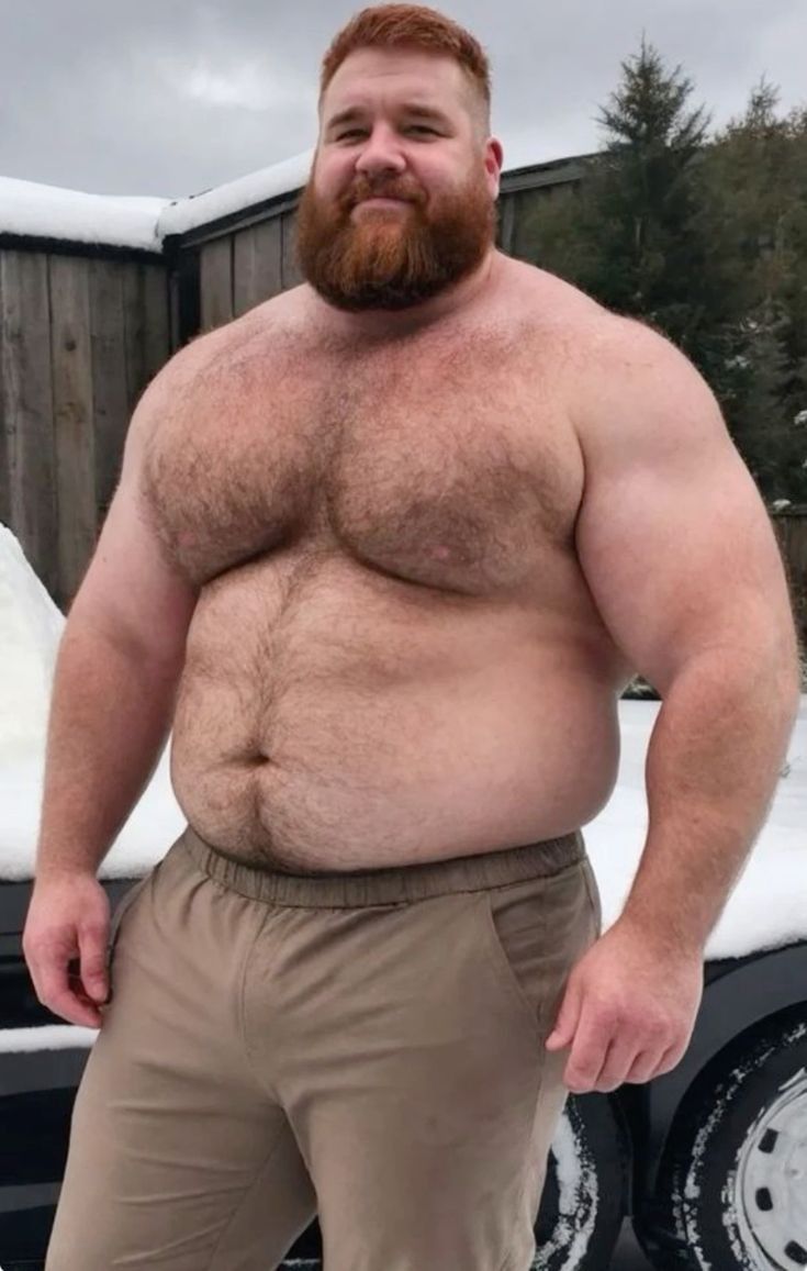 a muscular, shirtless redhead man with a beard stands outdoors in a snowy environment, wearing only tan pants.