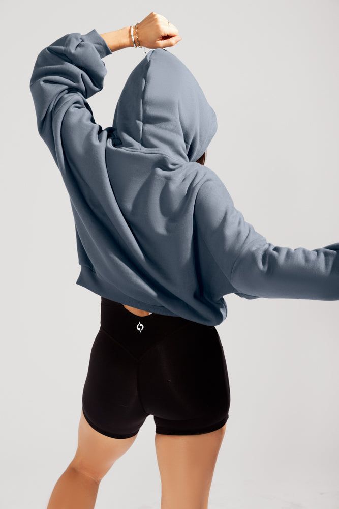 a woman wearing a blue cropped hoodie and black shorts strikes a dynamic pose with her arm raised, showcasing activewear in a studio setting.