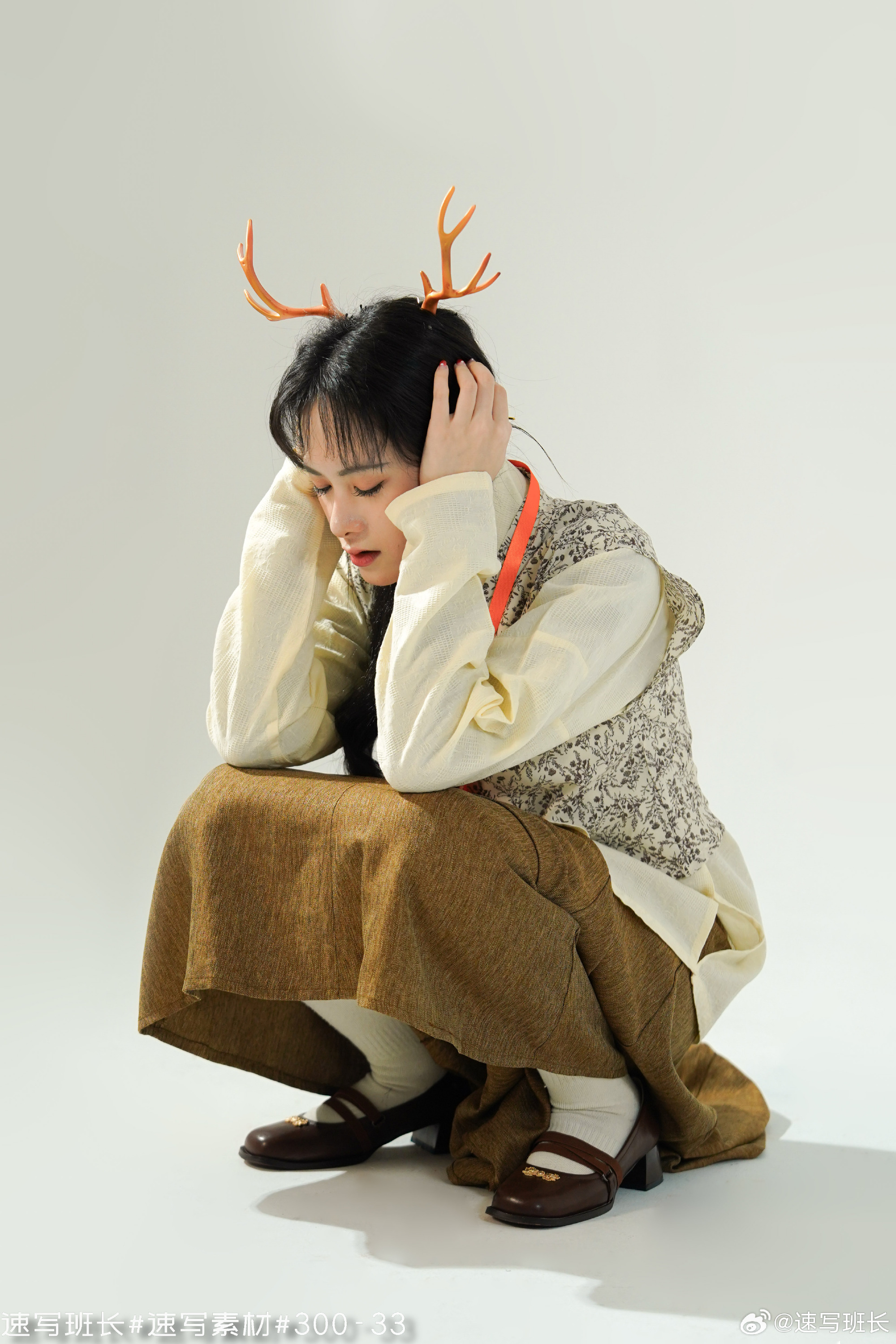 a young woman wearing a stylish outfit, including a white top, brown skirt, white socks, and brown shoes. she is crouching on the floor with her head down, her hands on her ears, and wearing a whimsical antler headband. the background is a bright white, creating a clean and minimalist aesthetic. the image suggests a sense of contemplation or perhaps a playful holiday spirit.