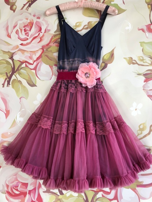 a sleeveless dress with a navy blue bodice and a deep v neckline. it has a multi layered, flowy skirt with ruffles and lace detailing. the skirt is a deep pink color. the dress has a velvet ribbon belt around the waist. there is a flower accent on the belt. the background of the image is floral.