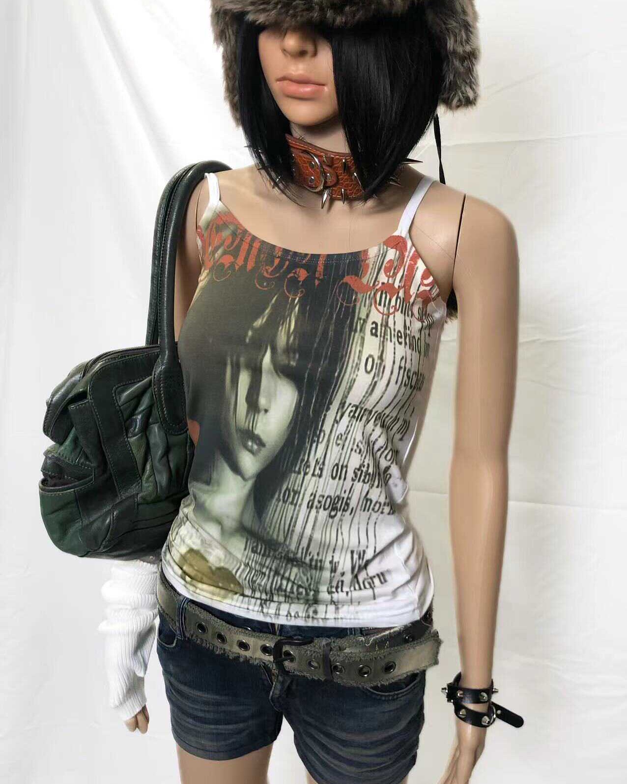 a mannequin dressed in alternative fashion poses against a white background.  she is wearing a white graphic tank top featuring a woman's face and text, distressed denim shorts, a studded belt, a green leather handbag, a trapper hat, a studded choker, and black wristbands. the overall style is dark, edgy, and gothic. the image is professionally styled for fashion photography.