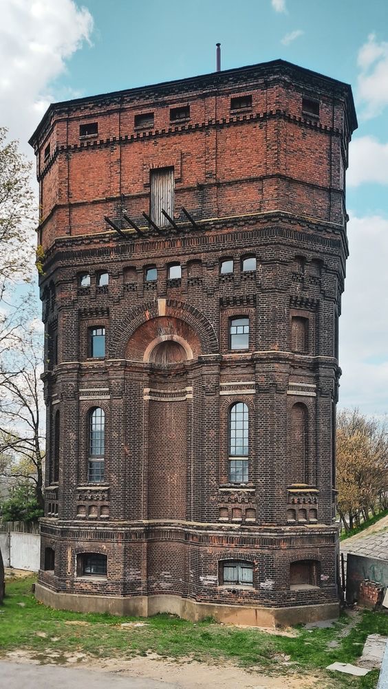 this is a photo of a tall brick tower with multiple stories and windows. it's an overcast day and the tower is set against a blue sky with clouds.  the tower has an old, weathered appearance, and there is grass and a dirt road in the foreground.