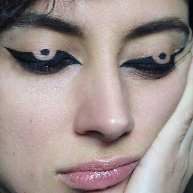 a closeup shot of a woman's face showcasing unique eyeliner makeup with a dot and wing design on both eyes. her skin has a natural tone, and she is resting her face on her hand.