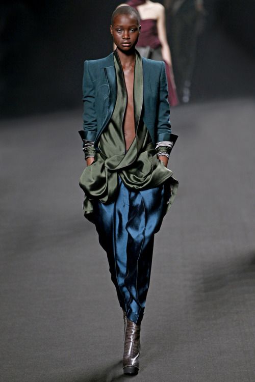a female model walks the runway during a fashion show. she's wearing a dark green blazer, a long silky green and blue dress, and black shoes. the woman is posing and her  hair is pulled back. the backdrop is a dark, blurred background.