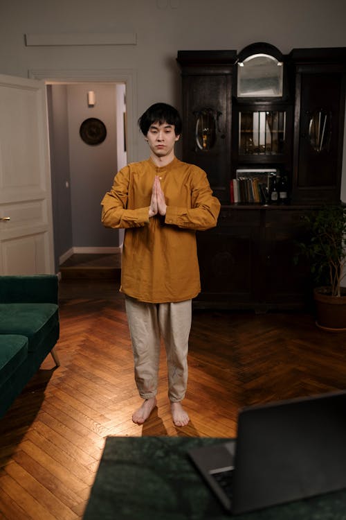 a young man stands in a living room, practicing a yoga or meditation pose with his hands together in prayer position. he's wearing a mustardyellow longsleeved shirt and light beige pants, barefoot on a polished wooden floor.  a laptop is partially visible in the foreground, suggesting he might be participating in an online class or session. the background includes a dark wooden cabinet and a partially visible doorway. the overall ambiance is peaceful and serene.