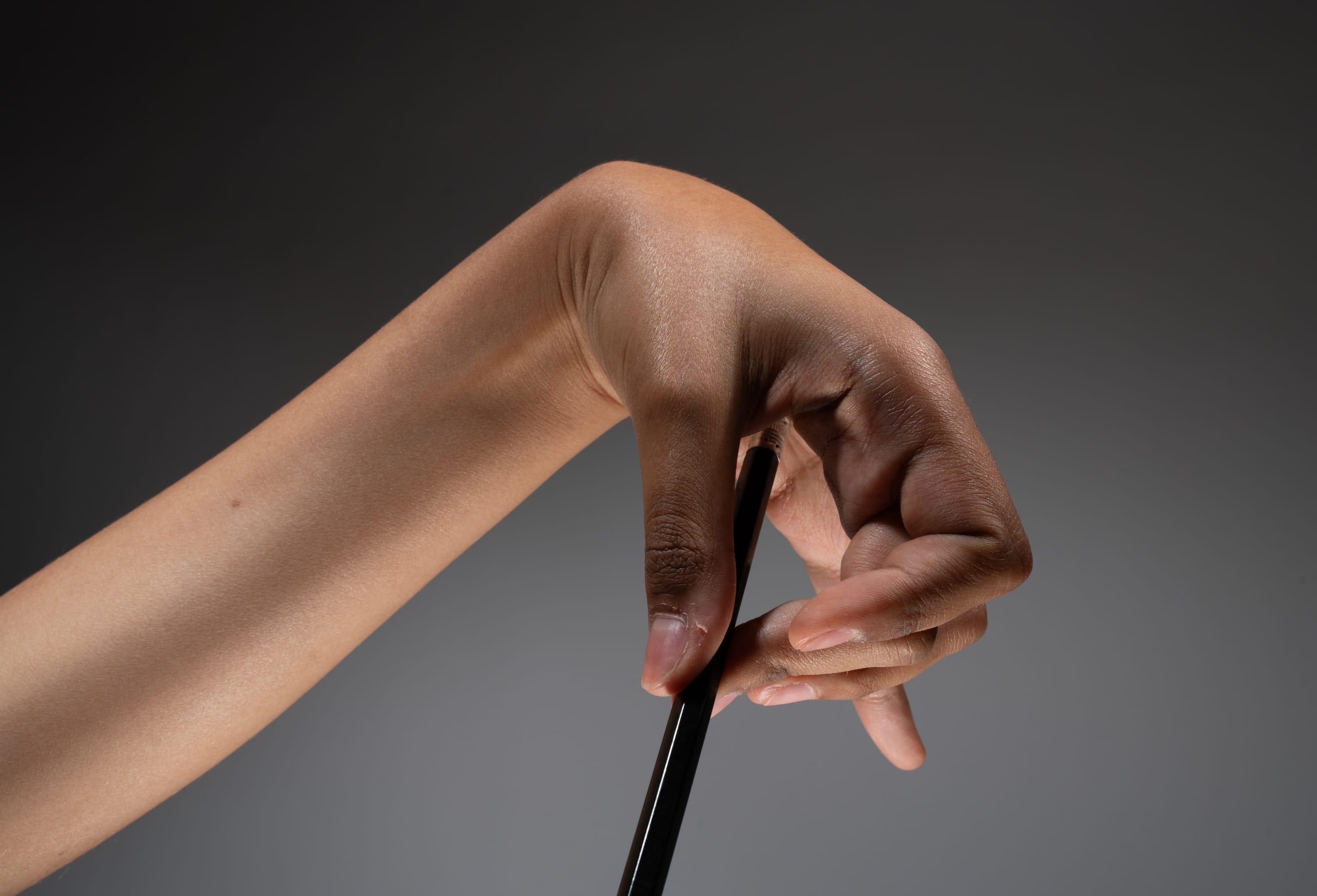 a closeup image of a hand holding a dark stick. the hand is positioned with the palm facing the viewer, and the stick is held between the thumb and index finger. the fingers are slightly curled around the stick, and the hand has a relaxed appearance. the arm is extended, and the skin has a smooth, even tone. the background is a neutral gray color, which helps to focus attention on the hand and stick. the lighting is soft and diffused, creating a naturallooking image.