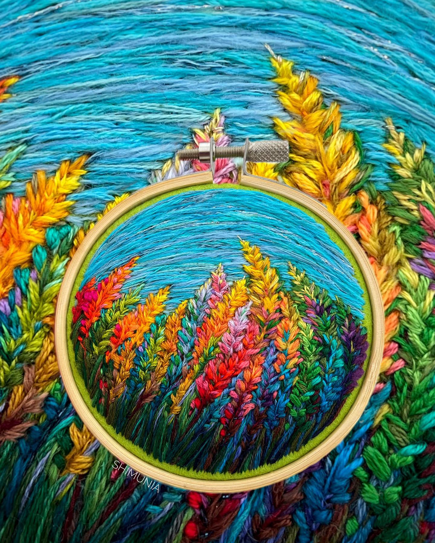 the image showcases a vibrant embroidery hoop art piece. set against a backdrop of a partially completed embroidery of a similar nature scene, the hoop displays a captivating landscape. lush green stitches form the base, from which sprout a colorful array of wildflowers in hues of yellow, orange, pink, and purple. the flowers reach towards the sky, stitched in various shades of blue, mirroring the water scene in the background. the artist's tag, "shimunia," is subtly incorporated into the greenery at the bottom of the hoop.