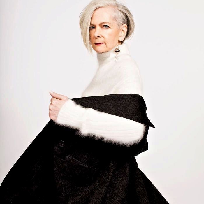 an elegant older woman with short gray hair is posing in a studio setting. she is wearing a white turtleneck sweater and a black and white patterned garment draped over her shoulders. she is also wearing dangle earrings. the background is a plain white.
