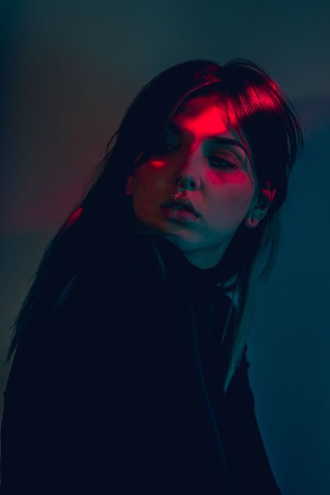a closeup portrait of a woman with long dark hair. her face is illuminated by dramatic red and blue lighting, creating a moody and evocative atmosphere.  the background is a dark teal.  the woman's expression is neutral, but the lighting adds a sense of mystery and intrigue.