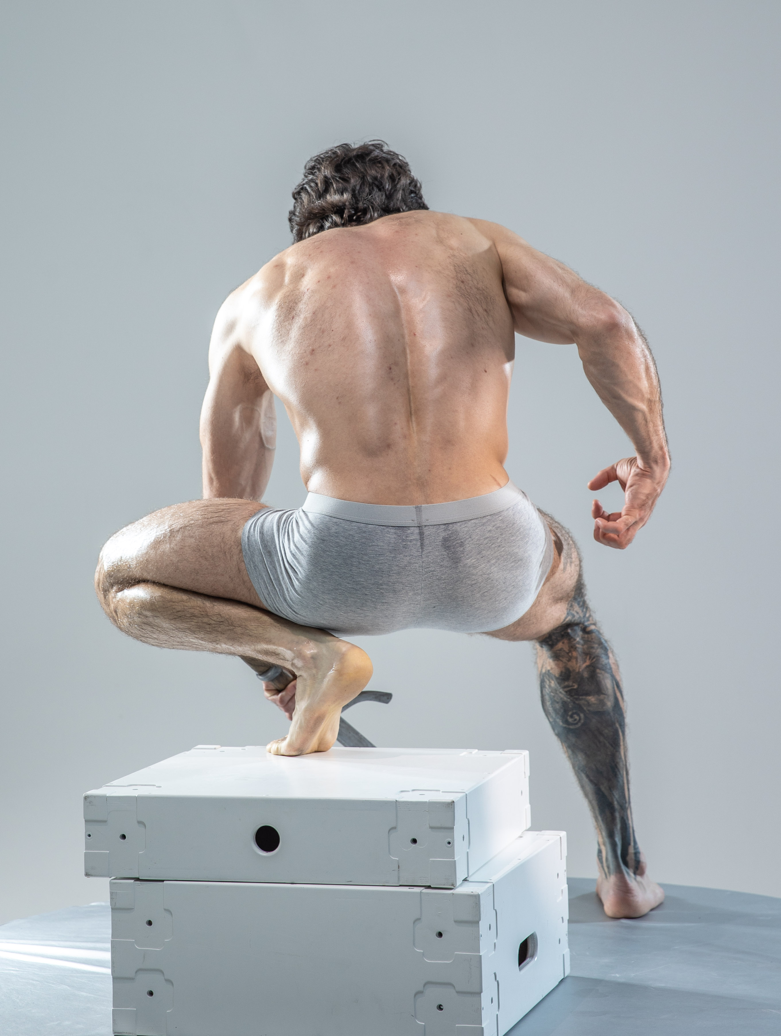 a muscular man with tattoos is captured from behind, showcasing his physique. he's wearing gray underwear and perched on the edge of two stacked white platforms, his bare feet gripping the surface. the studio setting is minimal with soft, even lighting.