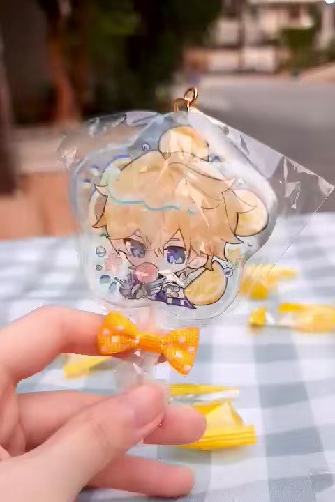 a hand is holding a keychain that is shaped like a lollipop.  it is made of clear plastic and has a picture of a young boy from an anime. the character has yellow hair, blue eyes, and is wearing a yellow and blue outfit. the lollipop is on a stick and is decorated with a yellow bow.  the keychain is on a checkered white and yellow background.  several other plastic wrapped yellow candy lollipops are visible in the background.