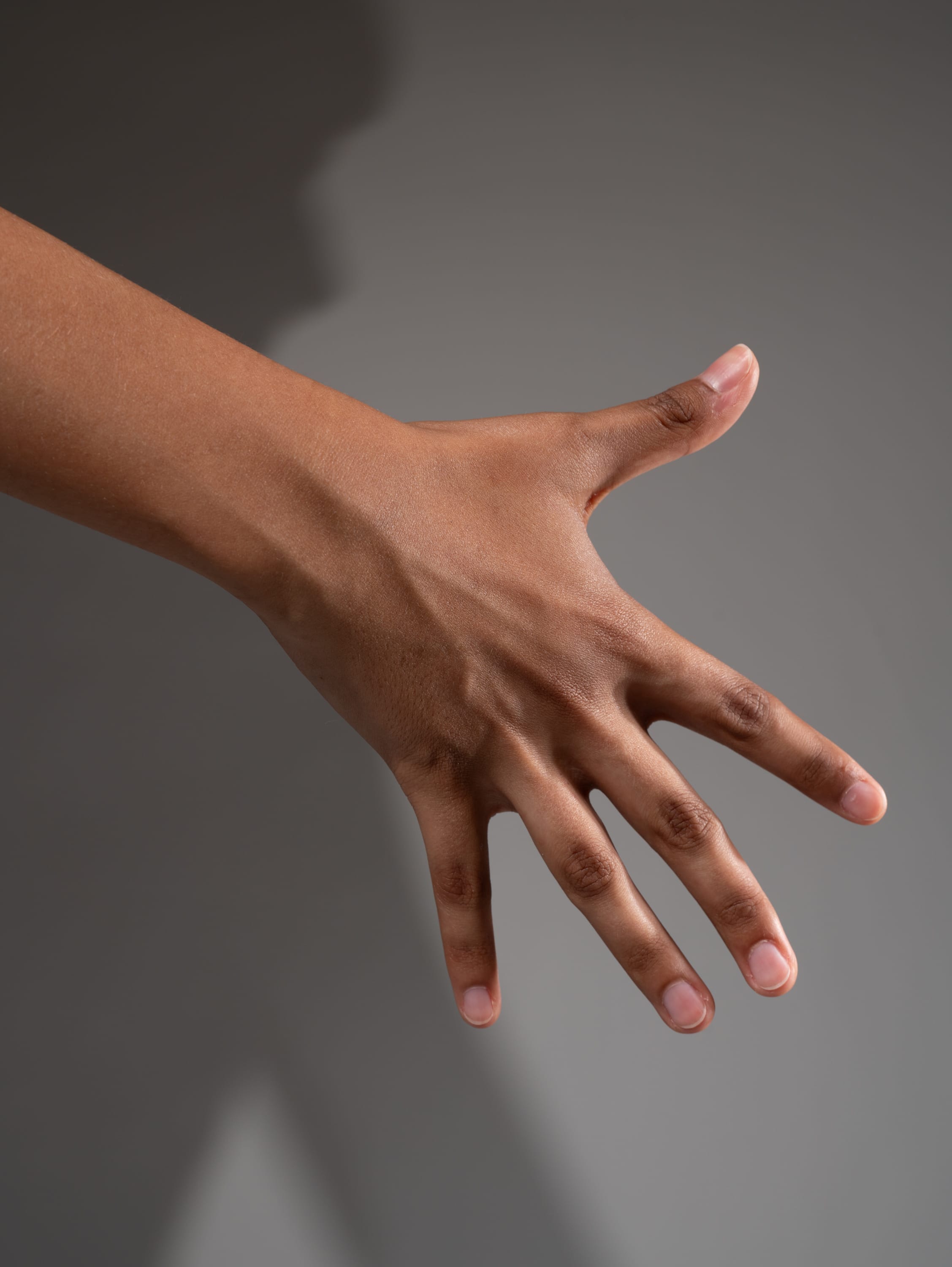 a closeup shot of a hand with brown skin extended against a gray background. the hand is positioned with the palm facing the viewer, and all five fingers are slightly spread apart.  a distinct shadow of the hand is cast onto the background, suggesting strong directional studio lighting.