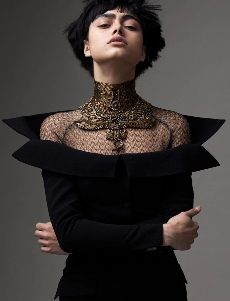 a woman with short black hair and light skin is posed against a gray background. she is wearing a black longsleeved dress with a wide, offtheshoulder collar. over a sheer, netlike layer covering her upper chest and neck, she wears an ornate gold collar necklace with an egyptianinspired design. her arms are crossed, and she looks upwards with a stoic expression. the image is a studio shot with a focus on high fashion and dramatic styling.