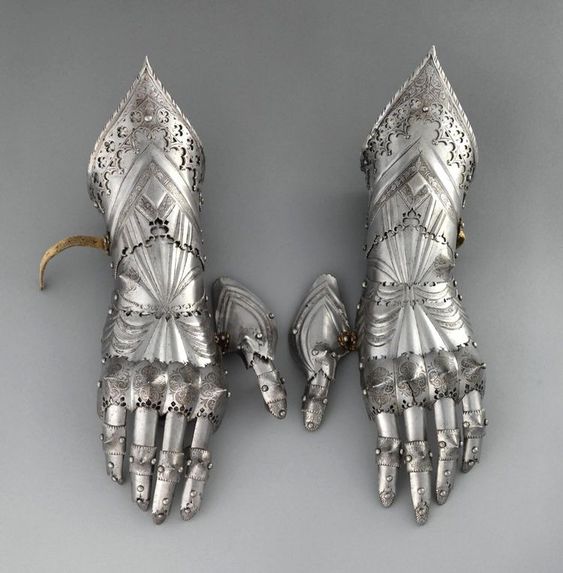 a pair of ornate, intricately crafted metal gauntlets from a museum collection. the gauntlets are made of steel with a polished finish, showcasing a high level of craftsmanship.  they feature articulated fingers for flexibility and are adorned with decorative engravings.  two additional pieces lie beside the gauntlets, likely offering alternative hand protection.