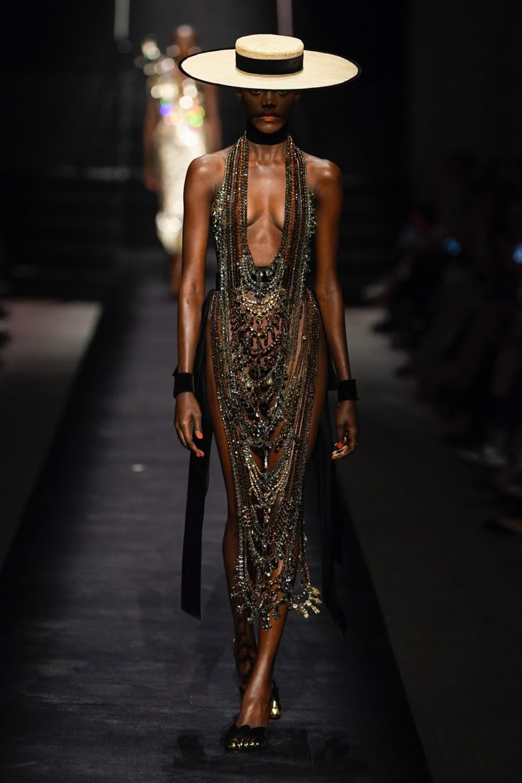 a model is walking down a runway during a fashion show. she is wearing a widebrimmed hat with a black band, a black choker, and a sheer, embellished dress with gold jewelry. the dress has a plunging neckline and is decorated with beads and other embellishments. she is wearing black shoes. she is wearing a black ribbon as wrist cuffs on both hands. the runway is black, and there are people in the background.