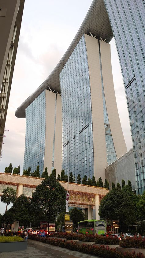 a lowangle view of the iconic marina bay sands hotel in singapore. the three towers are prominent, with their distinctive curved rooftop.  the image also captures the surrounding cityscape, including lush greenery, streets, and buses. the sky is partly cloudy, and the overall lighting suggests it's likely late afternoon or early evening.  the perspective emphasizes the height and grandeur of the building.