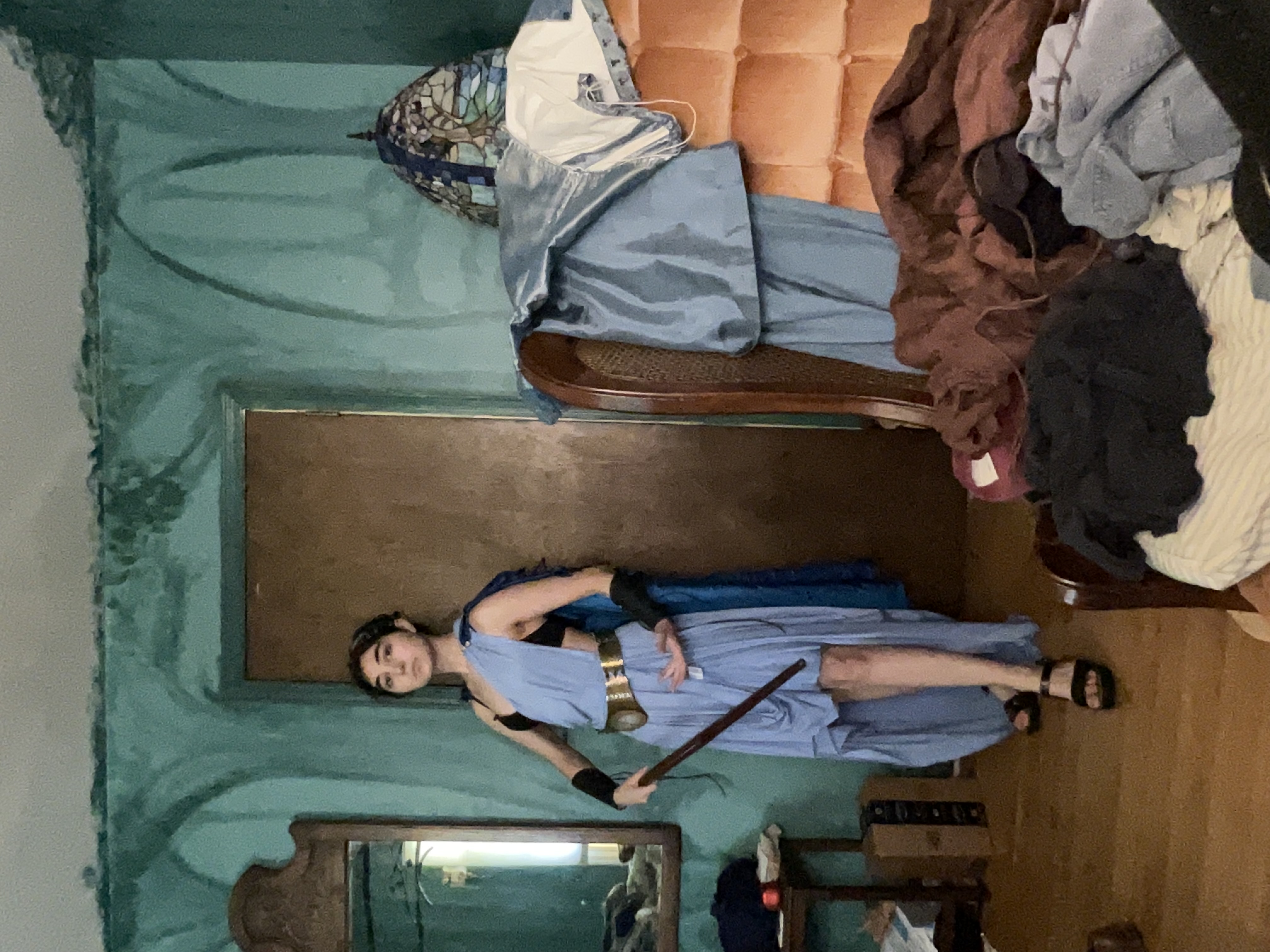 young woman in a blue greekstyle cosplay costume stands in a messy room.  she's wearing a light blue, asymmetrical dress, dark sandals, and armbands. she holds a long, dark staff.  the room's walls are teal and somewhat distressed, and the furniture and floor show signs of disorganization and clutter, with clothing piled on a chair and bed.  there's a mirror and a stainedglass lamp in the background.  the overall impression is one of casual, perhaps amateurish, cosplay in a livedin space.